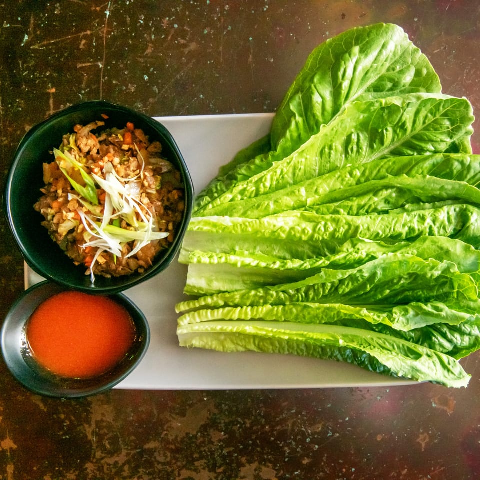 Lettuce wraps with a bowl of savory filling, garnished with scallions, and a side of red sauce.