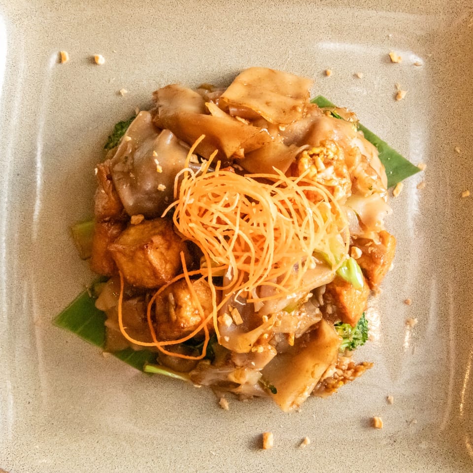 Pad See Ew with wide rice noodles, tofu, broccoli, and shredded carrots on a banana leaf.