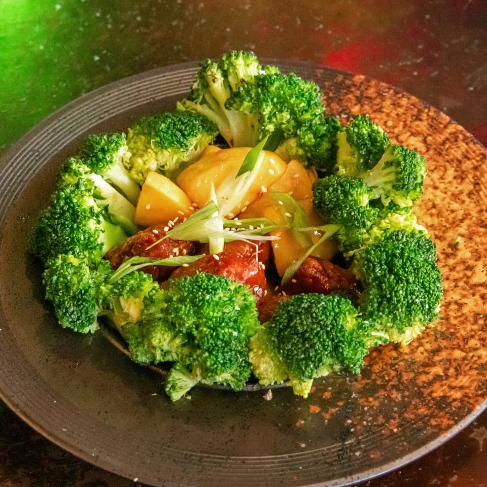 Sweet and sour chicken with pineapple, broccoli, scallions, and sesame seeds on a dark plate.