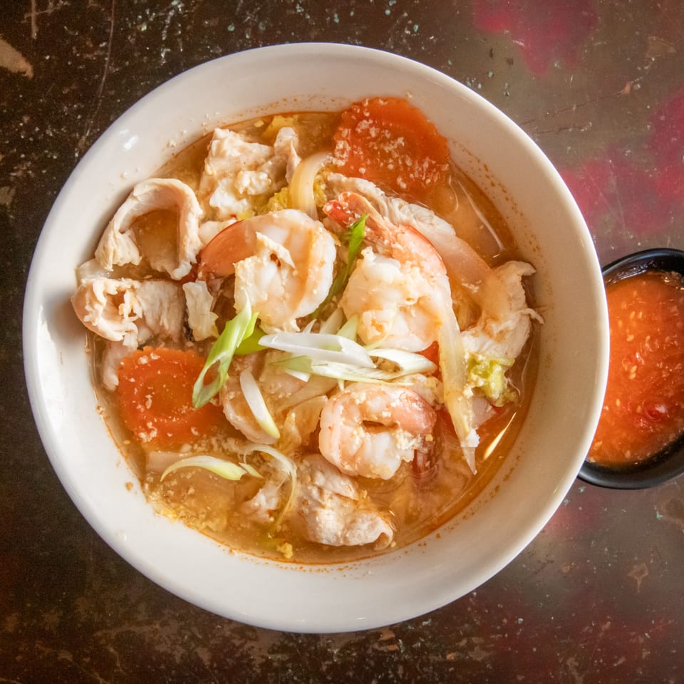A bowl of Tom Yum soup with shrimp, chicken, and vegetables, with a side of chili sauce.